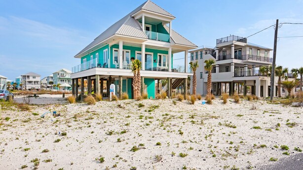 Een tweelaags huis aan het strand met een balkon, gelegen op een zandstrand met andere huizen op de achtergrond.