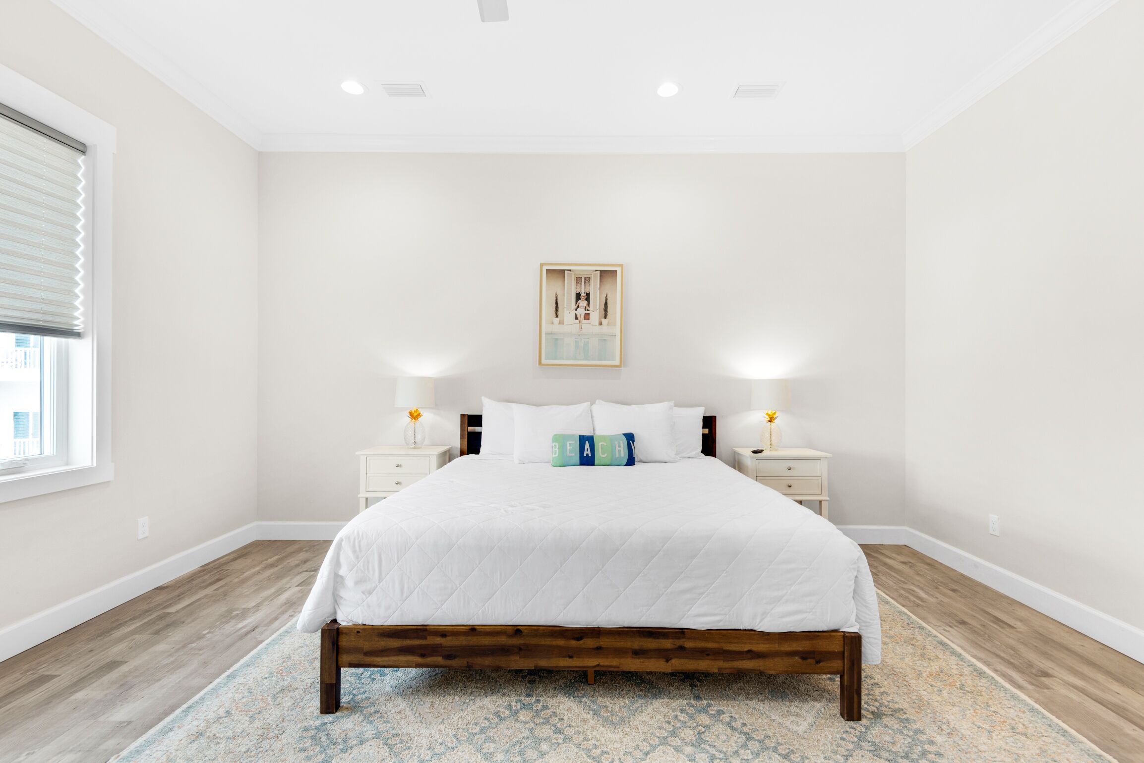A bedroom with a wooden bed frame, white bedding, two bedside tables with lamps, a framed picture on the wall, and a window with blinds.