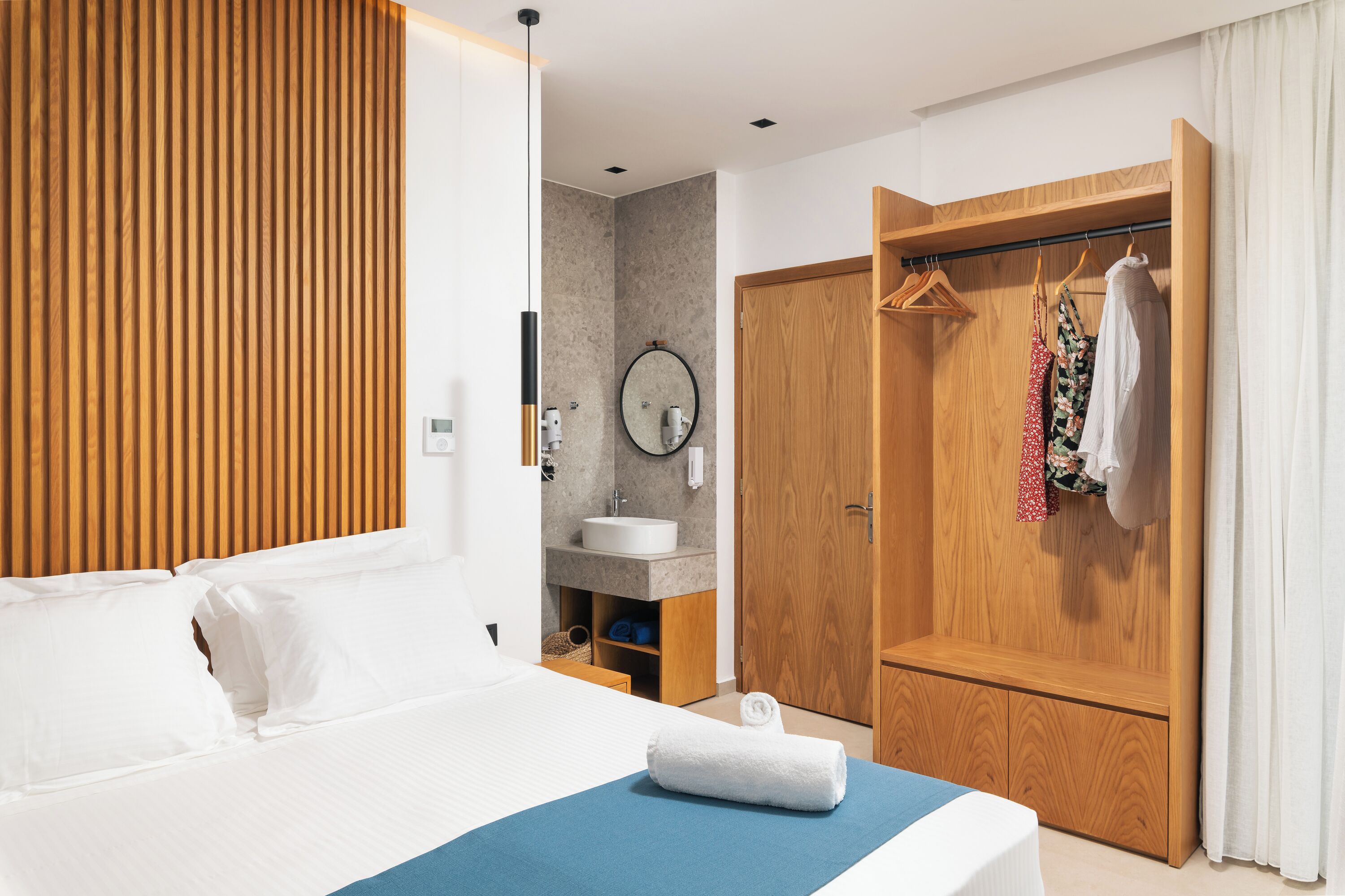 Une chambre d’hôtel avec un lit, une armoire en bois, une salle de bain avec un lavabo et un miroir, et une couvre-lit bleue.