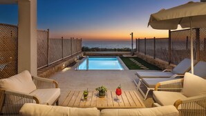 Área de piscina no terraço com espreguiçadeiras, uma mesa e vista para o pôr do sol sobre o oceano.