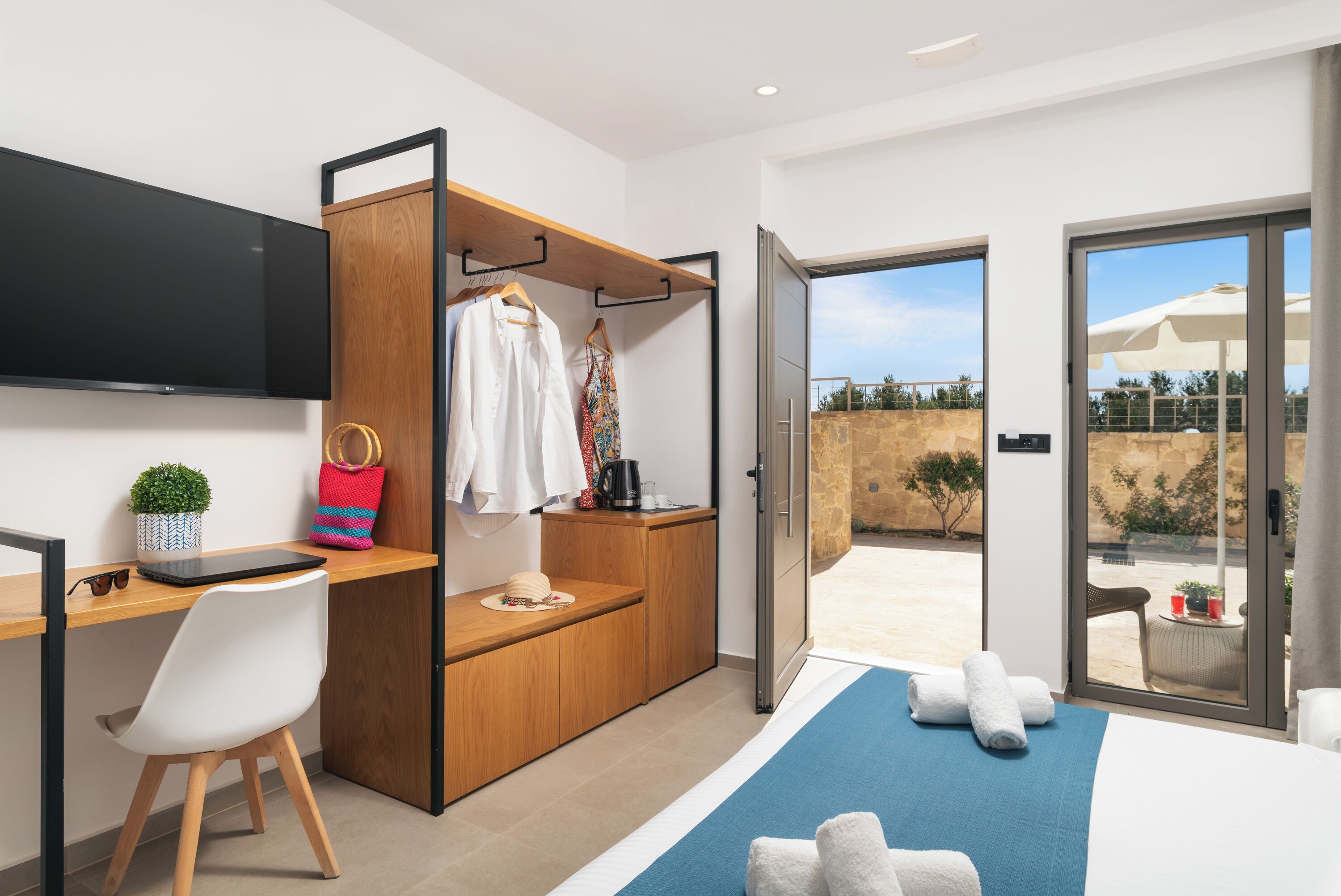 A modern hotel room with a wooden desk, a TV mounted on the wall, a wardrobe with a hanging rack, and a large sliding glass door leading to an outdoor area.