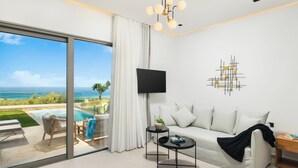 Uma sala de estar moderna com um sofá, uma mesa de centro e uma televisão. Há uma janela ampla com vista para o oceano e uma piscina.