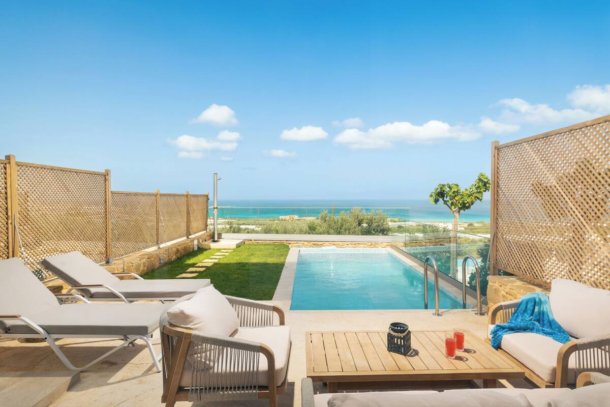 Ein Poolbereich mit Liegestühlen, einem Holztisch und Blick aufs Meer.