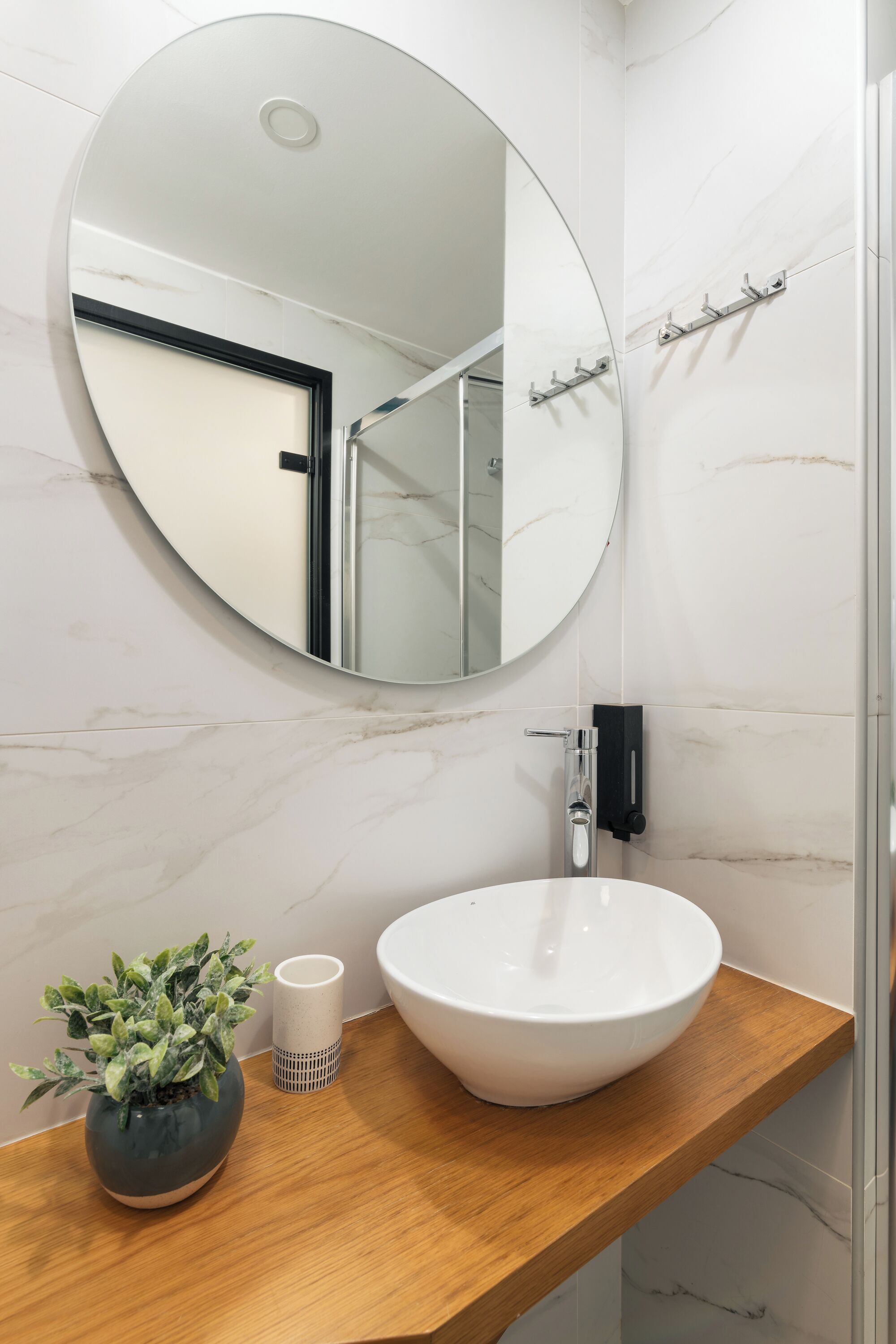 A bathroom with a round mirror, a white sink, a wooden countertop, a potted plant, and a white cup.