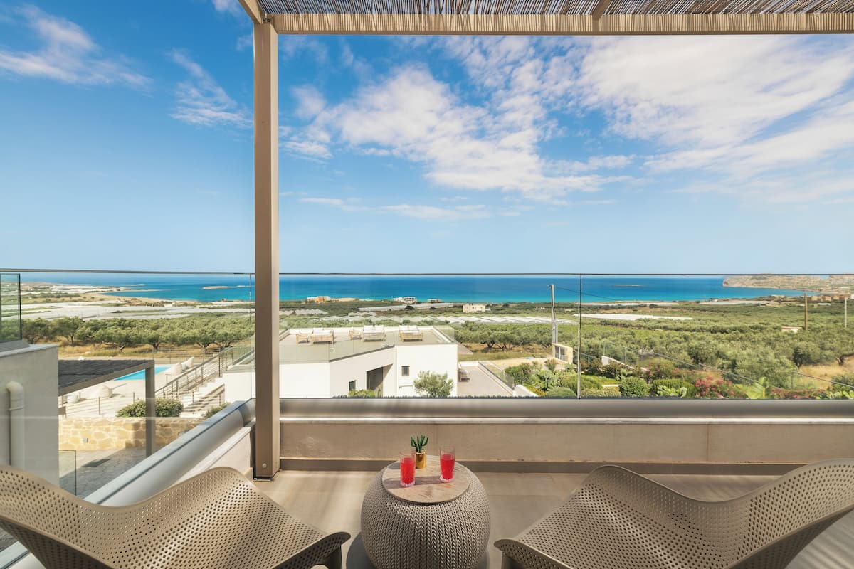 Ein Balkon mit zwei Korbstühlen, einem kleinen runden Tisch und einem Blumentopf. Der Blick öffnet sich über eine Küstenlandschaft mit Gebäuden und viel Grün.