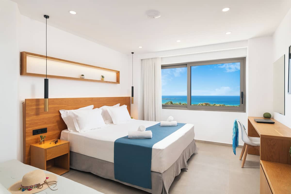 Ein modernes Hotelzimmer mit einem großen Bett, einem Schreibtisch, einem Sessel und einem Nachttisch, mit Blick aufs Meer.