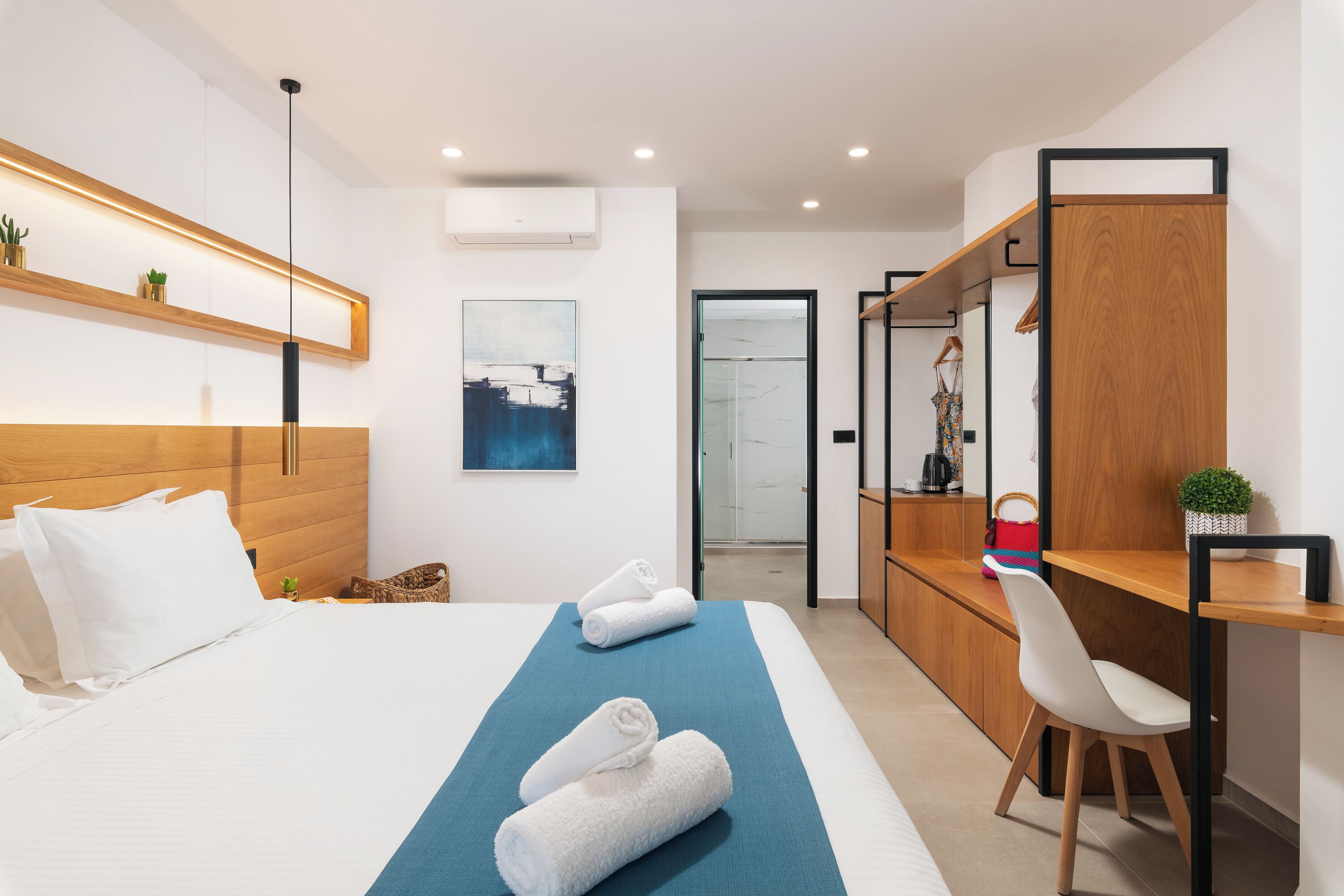 Une chambre d’hôtel moderne avec une tête de lit en bois, un lit recouvert de draps blancs, un tapis bleu décoratif, une chaise blanche, un bureau en bois et une porte de douche en verre.