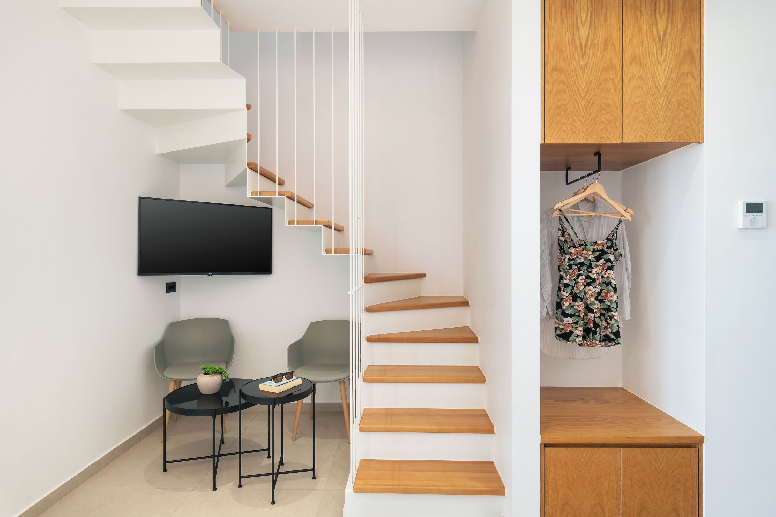 A modern living space with a staircase, a flat-screen TV, two chairs, a small table with a plant, and a wooden cabinet.