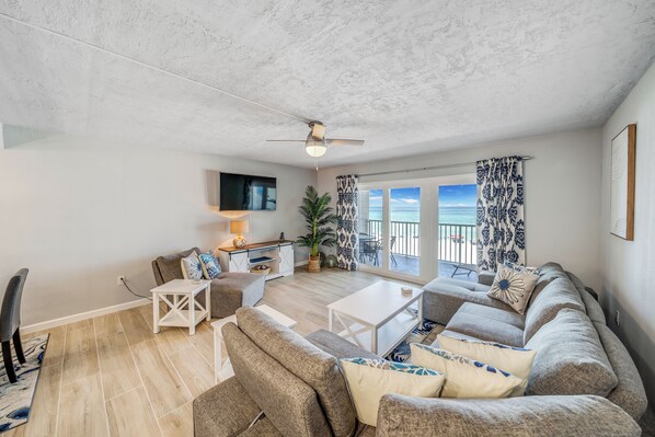 A spacious living room with a ceiling fan, a flat-screen TV, a dining area, and a large window with a view of the outdoors.