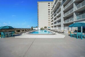 A swimming pool surrounded by lounge chairs and umbrellas, with a tall building in the background. - FREE Golf & Perks! Updated Mariner West 406, Swimming Pool, Tennis & Grills! (Panama City Beach)