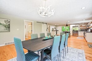 A spacious dining area with a dark wooden table and light blue upholstered chairs, a chandelier, and a view of the kitchen in the background.