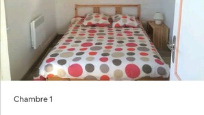 A small, single-bed room with a polka dot bedspread, a wooden headboard, a bedside table with a lamp, and a white radiator. - Appartement 8/10 Personnes Face à la mer (Barbâtre)