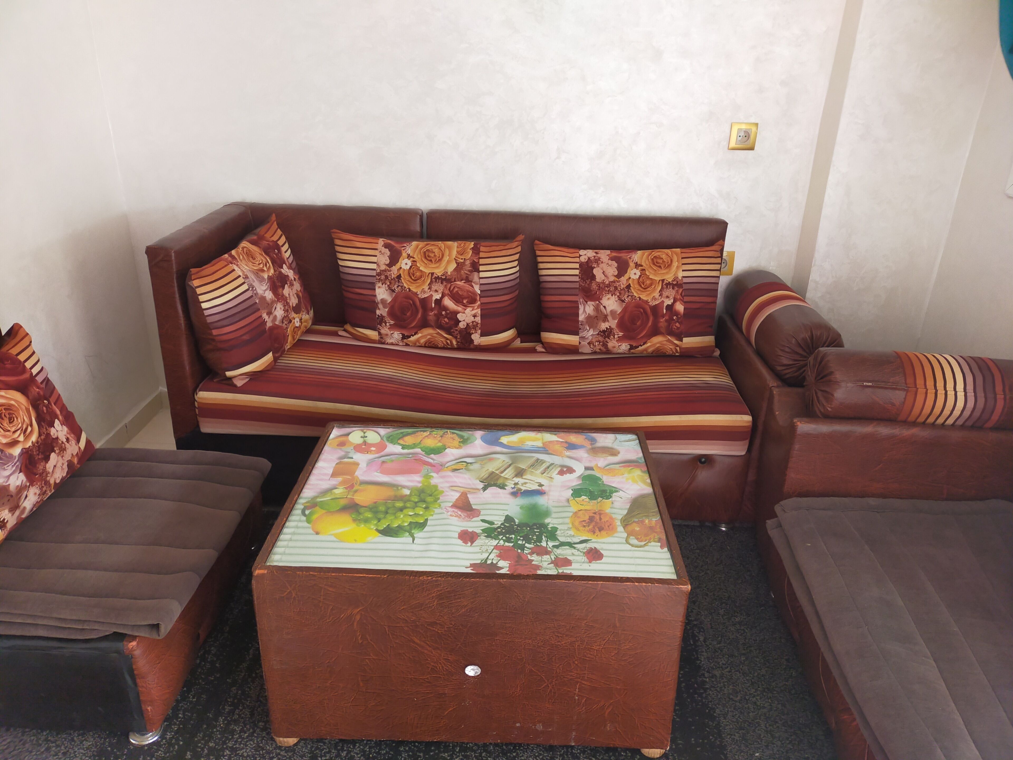 A living room with a brown leather sofa, striped cushions, and a floral-patterned throw. A small wooden coffee table with a fruit design is in front of the sofa.
