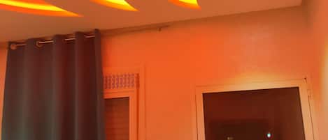 A room with a modern ceiling design featuring illuminated wave patterns. There is a window with a view of the outside, a door, and a curtain partially drawn.
