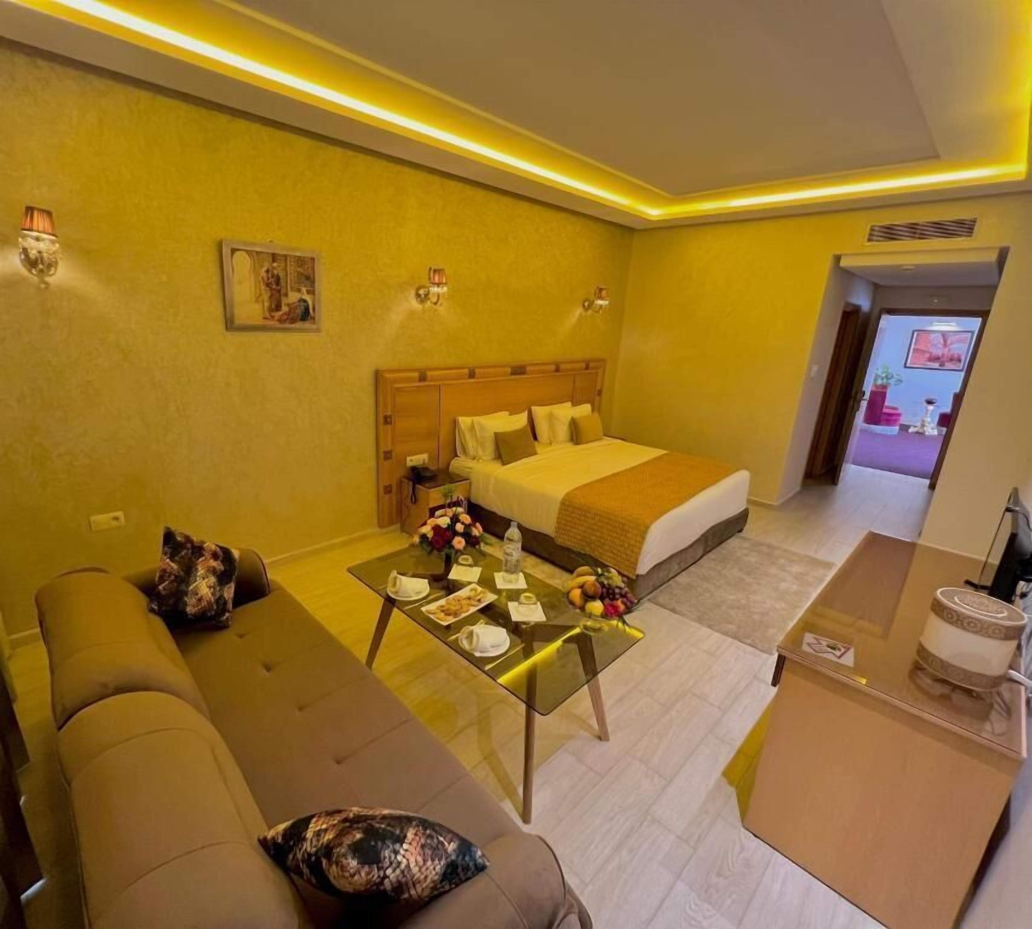 Una habitación de hotel con una cama, un sofá, una mesa baja con frutas y una mesita de noche con una lámpara.