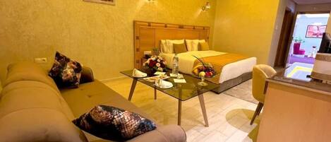 Habitación de hotel con cama, sofá, mesa de centro de vidrio y frutero, y un escritorio con lámpara.