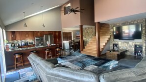 Uma sala de estar ampla com um sofá seccional em forma de L, cozinha com armários de madeira, área de jantar e parede de destaque em pedra com uma TV de tela plana.