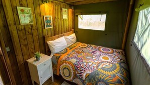 A cozy room with a wooden wall, a bed with a colorful patterned blanket, a small bedside table with a plant, and a window with a view of greenery. - Awe Inspiring two storey tent (Crowhurst)