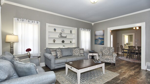 Un salon avec un canapé gris, une table basse et des fauteuils à motifs. On y trouve une étagère blanche ornée d’objets décoratifs et un tableau accroché au mur.