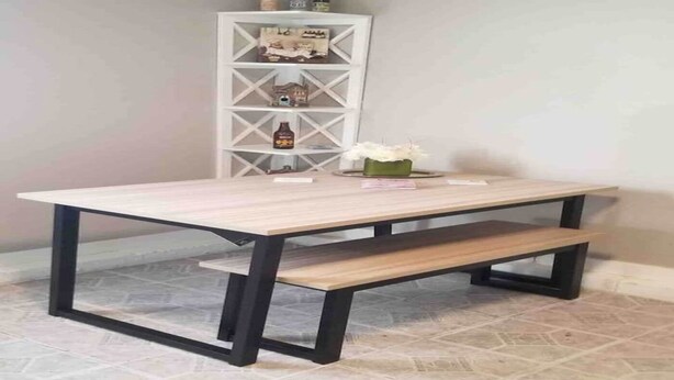 Une table rectangulaire avec un plateau en bois clair et des pieds en métal foncé, accompagnée d’un banc assorti. Une étagère blanche avec des portes en verre est placée derrière la table et expose divers objets.