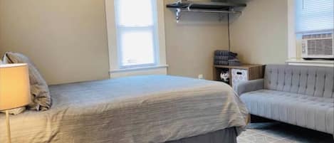 A hotel room with a bed, a sofa, a TV mounted on the wall, and a window with blinds.