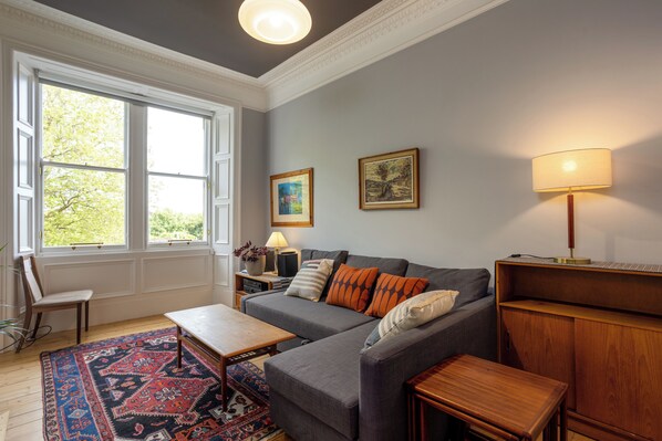 A living room with a grey sofa, a wooden coffee table, a patterned rug, a side table with a lamp, and a window with a view of greenery.