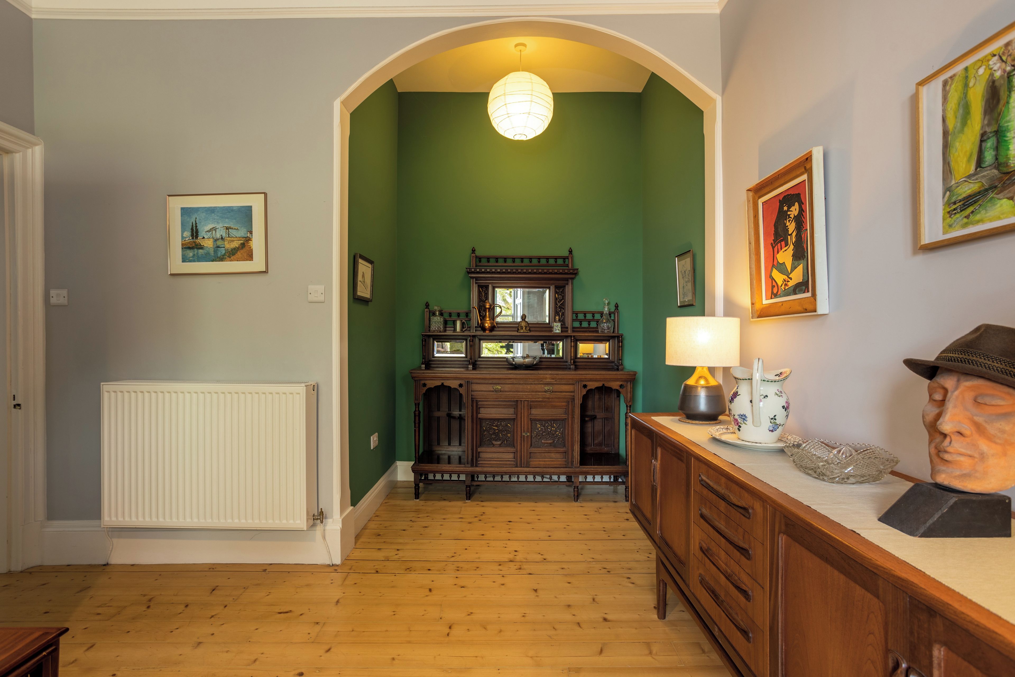 Un couloir avec un radiateur, un plancher en bois, des murs verts, un miroir et des œuvres d’art.