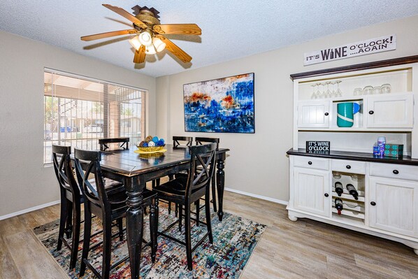 A dining area with a wooden table and chairs, a ceiling fan, a large abstract painting, and a wine cabinet with glassware.