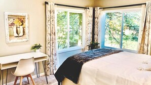 Ein Schlafzimmer mit einem Bett, einem Schreibtisch und einem Stuhl. Zwei Fenster mit Vorhängen sind vorhanden.