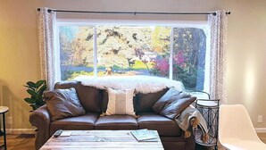Ein Wohnzimmer mit braunem Ledersofa, einem weißen Fellüberwurf und Blick auf einen Garten mit Bäumen und Blumen.