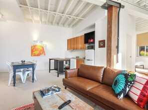 A living room with a brown leather sofa, a coffee table with books and a remote, a dining area with a table and chairs, and a kitchen with wooden cabinets and appliances. - Heritage Woolstore Apartment | Teneriffe, Brisbane (Teneriffe)