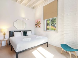 A modern bedroom with a bed, bedside table, lamp, and a window with a view of greenery.