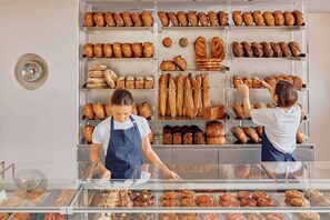 A bakery with shelves of bread and pastries, a display case with baked goods, and two individuals in aprons. - Heritage Woolstore Apartment | Teneriffe, Brisbane (Teneriffe)