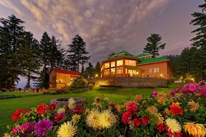 Une maison à deux étages, dotée de fondations en pierre et d’un toit végétalisé, entourée d’un jardin bien entretenu où s’épanouissent des fleurs aux couleurs vives.
