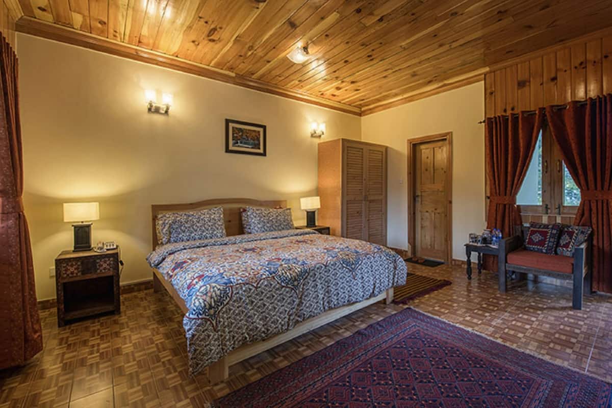 A bedroom with a wooden ceiling, a large bed with patterned bedding, two bedside lamps, a nightstand, a wooden wardrobe, and a red carpet.
