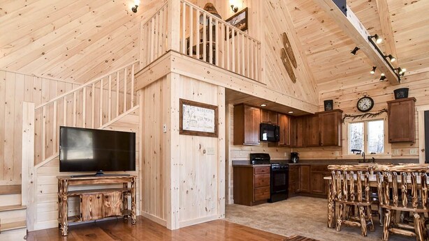 A spacious wooden cabin interior with a TV, wooden furniture, a kitchen, and a staircase. - Stay on Kingdom Trails in a four bedroom cabin! (Burke)