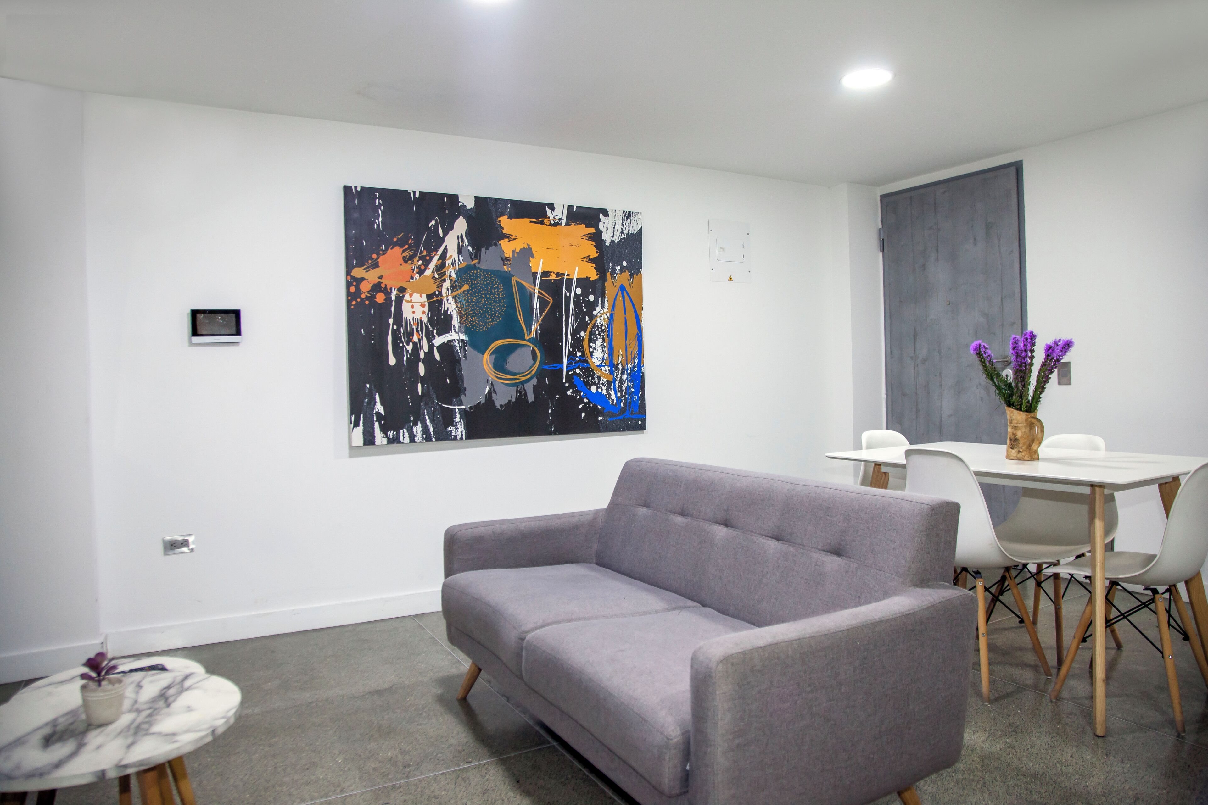 Ein modernes Wohnzimmer mit einem grauen Sofa, einem runden Tisch und einem Essbereich mit einem weißen Tisch und Stühlen. Ein großes abstraktes Gemälde hängt an der Wand, und im Hintergrund ist eine Tür zu sehen.