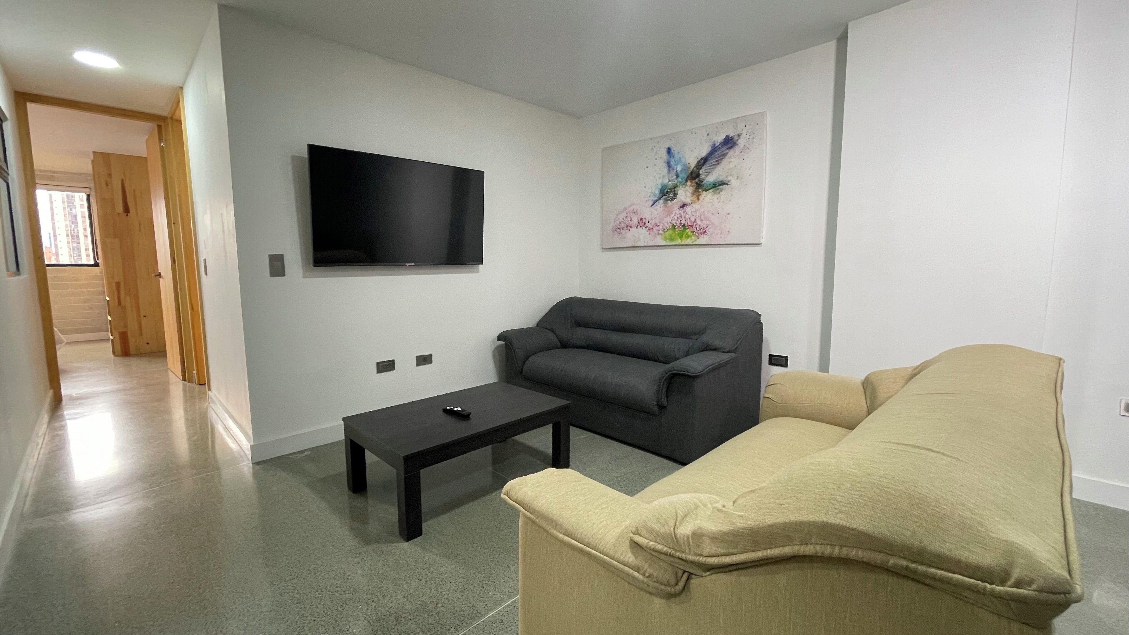 Un salon avec un téléviseur à écran plat fixé au mur, un canapé gris, un canapé beige et une petite table basse noire.