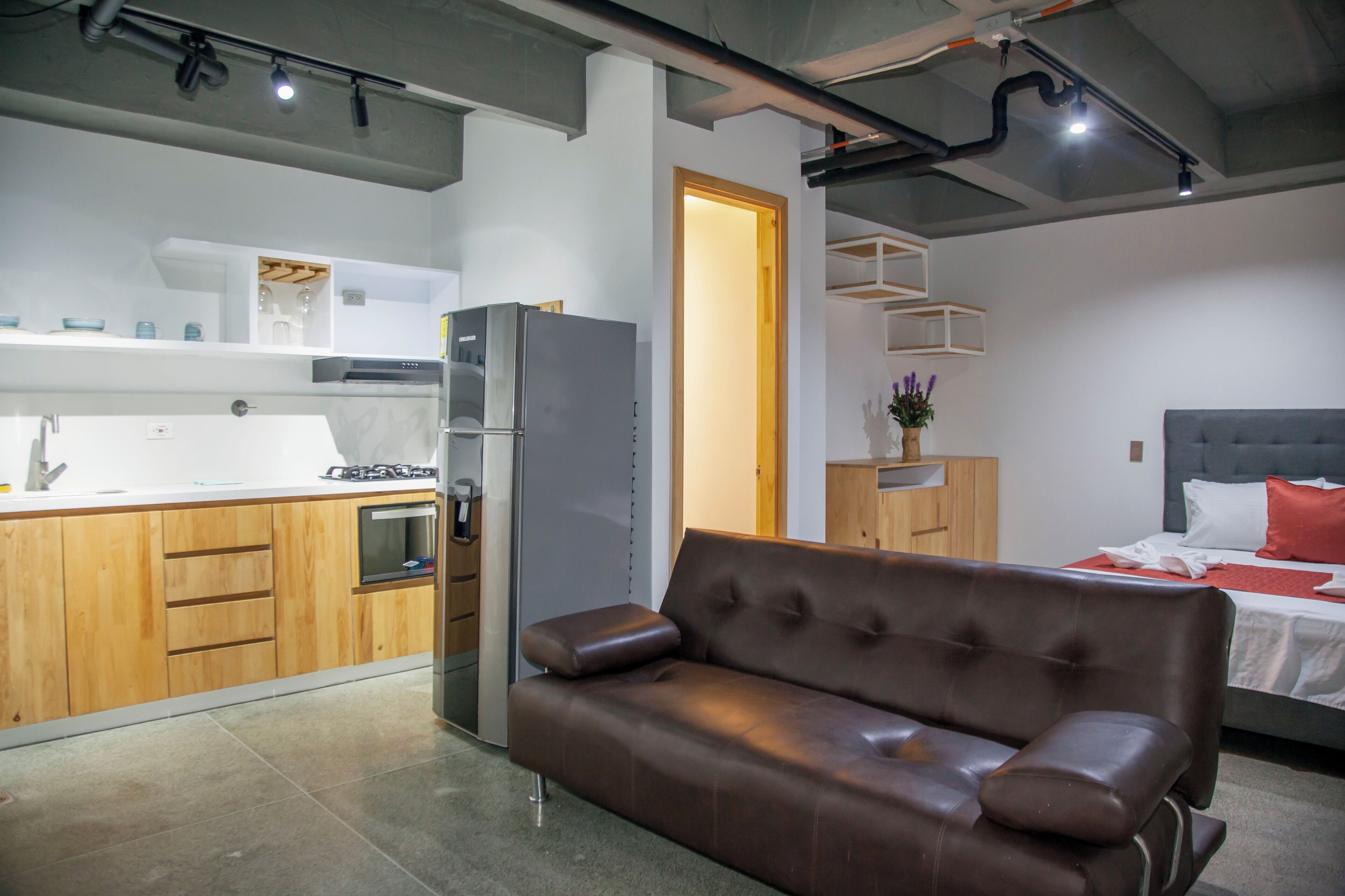A modern kitchen with a refrigerator, oven, and wooden cabinets. There is a brown leather sofa in the living area and a bed with a red pillow in the bedroom.