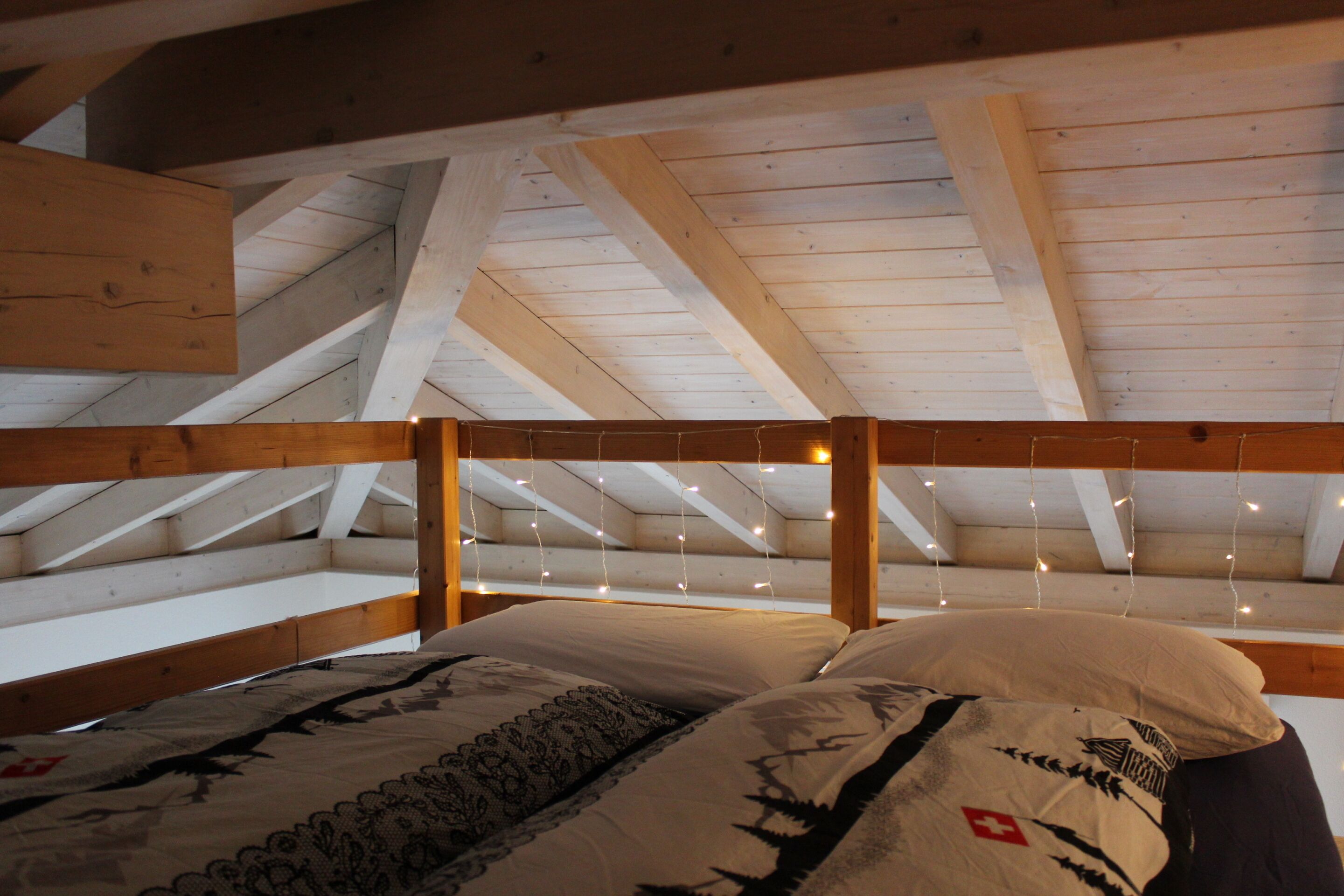 Dormitorio tipo loft con vigas de madera a la vista, techo inclinado y luces colgantes.