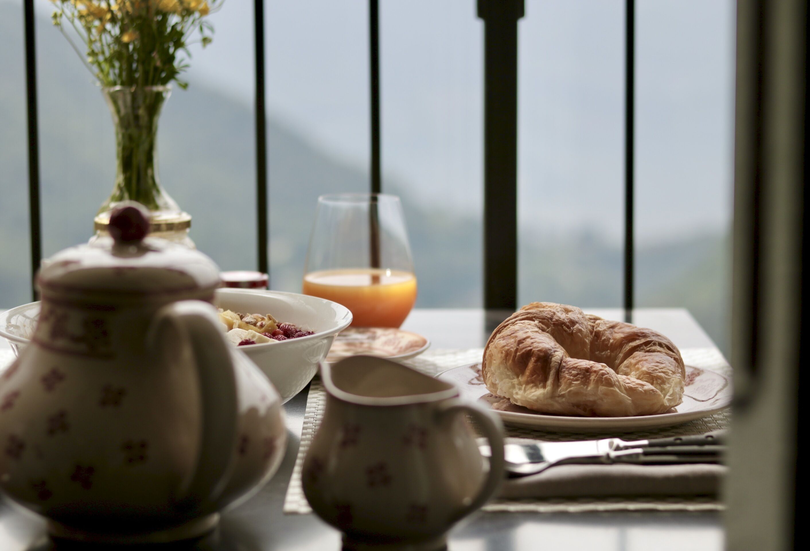 Ein Frühstückstisch mit Croissant, Teekanne, Tasse, Schale und Orangensaft, mit Blick auf Berge.