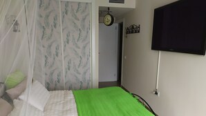 Un dormitorio con cama, una colcha verde, televisor en la pared, un reloj y una puerta que da a otra habitación.