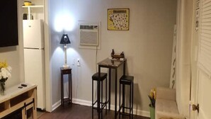 Un espace de vie compact comprenant un coin cuisine, un coin repas avec une table et des tabourets, une lampe et une affiche accrochée au mur.