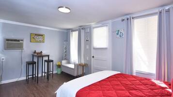 Un dormitorio con una colcha roja, un escritorio con dos sillas, una ventana con persianas y una puerta con un letrero.