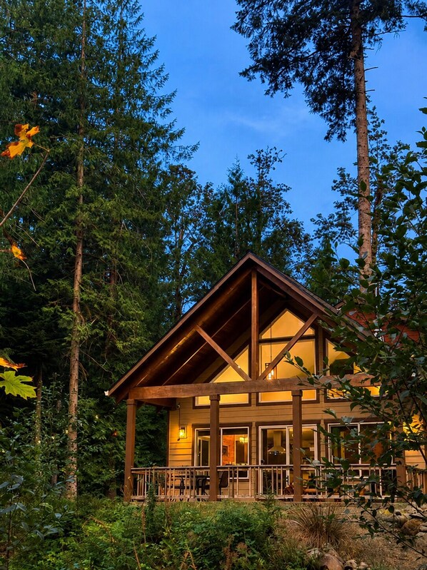 Une cabane avec une façade en bois, de grandes fenêtres et une véranda équipée d’un coin salon.