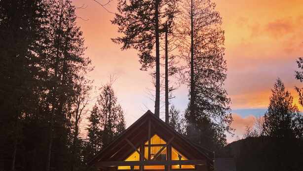 A cabin with a large front porch, multiple windows, and a sloped roof, surrounded by tall trees and set against a sunset sky. - Big Tree Retreat: Secluded cabin with hot tub, river views & woodstove (Welches)