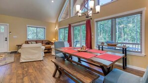Amplia sala de estar con una mesa de comedor de madera dispuesta para cuatro, ventanas grandes con cortinas rojas, un sofá y un banco.
