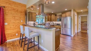 A spacious kitchen with wooden cabinets, a central island with bar stools, stainless steel appliances, and a large window providing natural light. - Big Tree Retreat: Secluded cabin with hot tub, river views & woodstove (Welches)