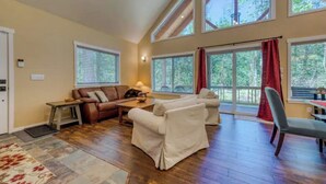 A spacious living room with a brown leather sofa, a beige armchair, and a wooden coffee table. Large windows provide natural light and a view of the outdoors. - Big Tree Retreat: Secluded cabin with hot tub, river views & woodstove (Welches)