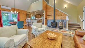 A spacious living area with a fireplace, wooden walls, and a kitchen in the background. - Big Tree Retreat: Secluded cabin with hot tub, river views & woodstove (Welches)